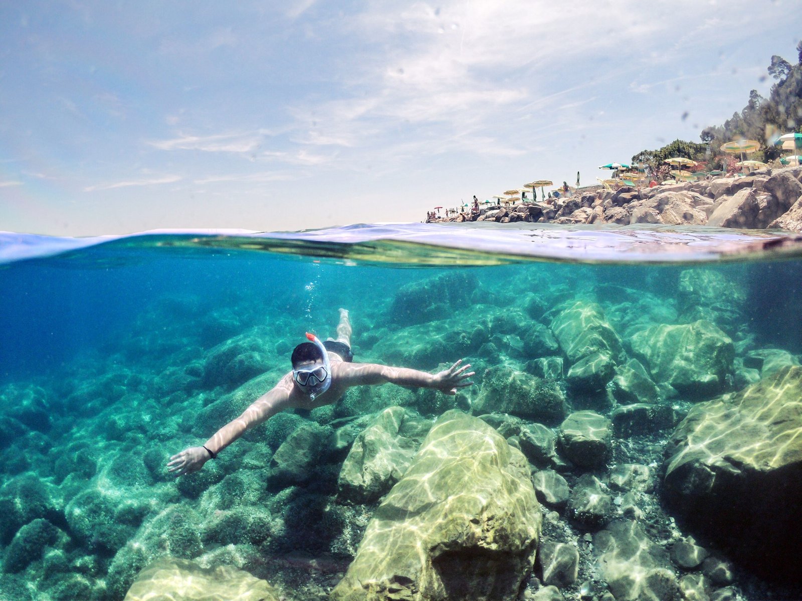 A snorkeler swims underwater near a rocky coastline under clear skies, showcasing the beauty of marine life and outdoor adventure.