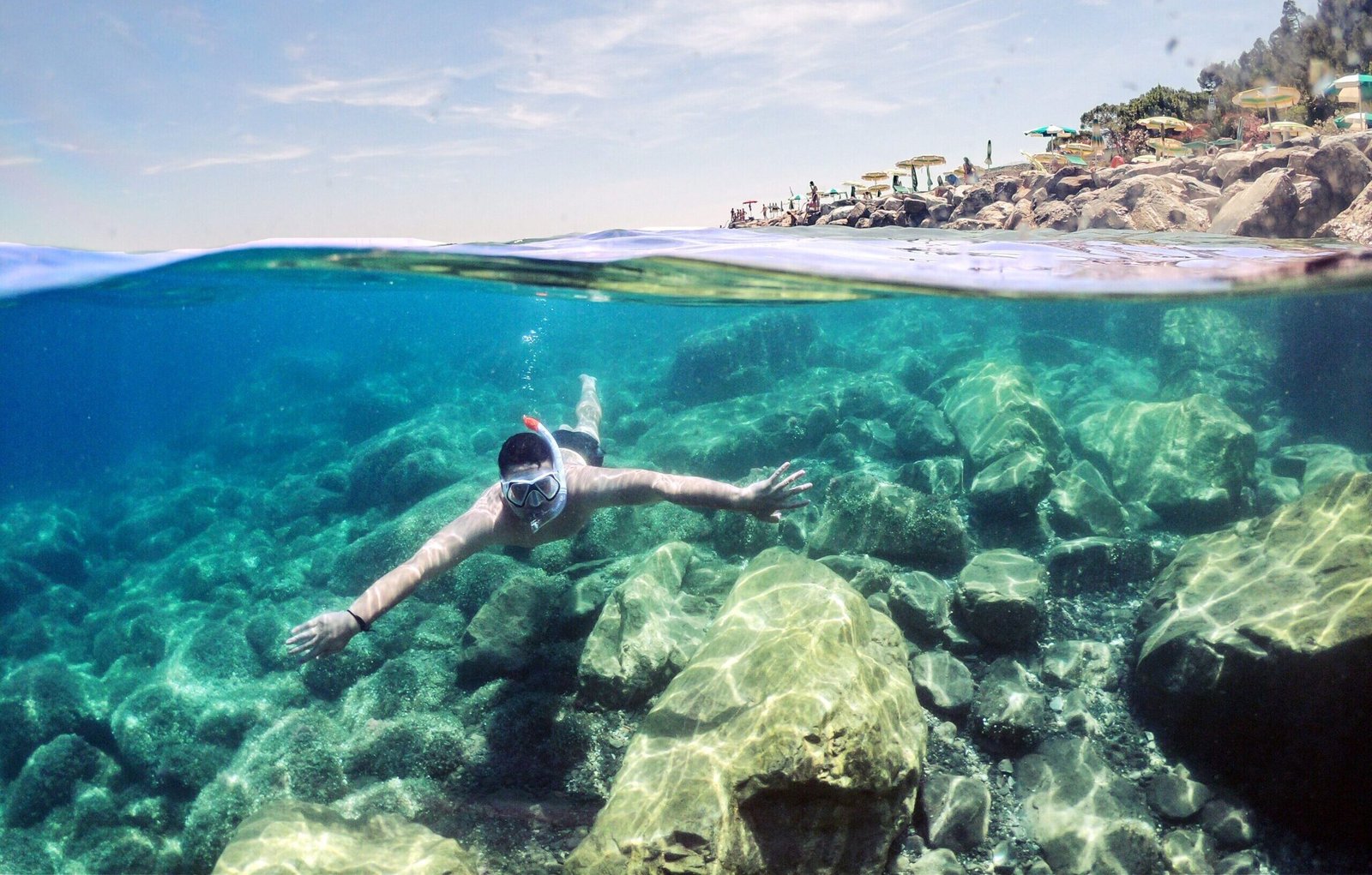 A snorkeler swims underwater near a rocky coastline under clear skies, showcasing the beauty of marine life and outdoor adventure.